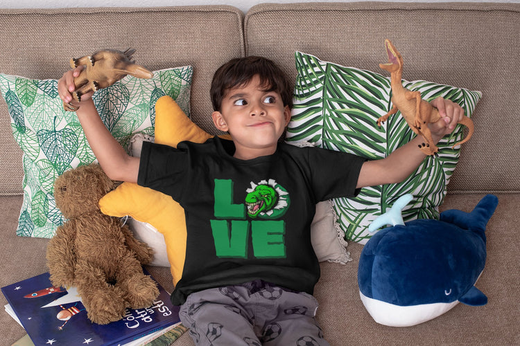 A young child holding toy dinosaurs in each hand wearing a t-shirt that has green letters spelling out the word LOVE but the letter O has a tyrannosaurus rex bursting through and out of it
