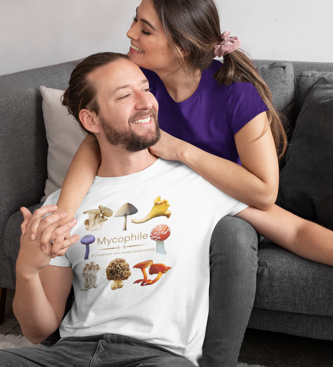 A man with a t-shirt saying 'mycophile - a person who loves mushrooms' with several real photos of mushrooms such as morel, chanterelle, etc. He is leaning against a woman in a t-shirt on a couch, both relaxed and smiling