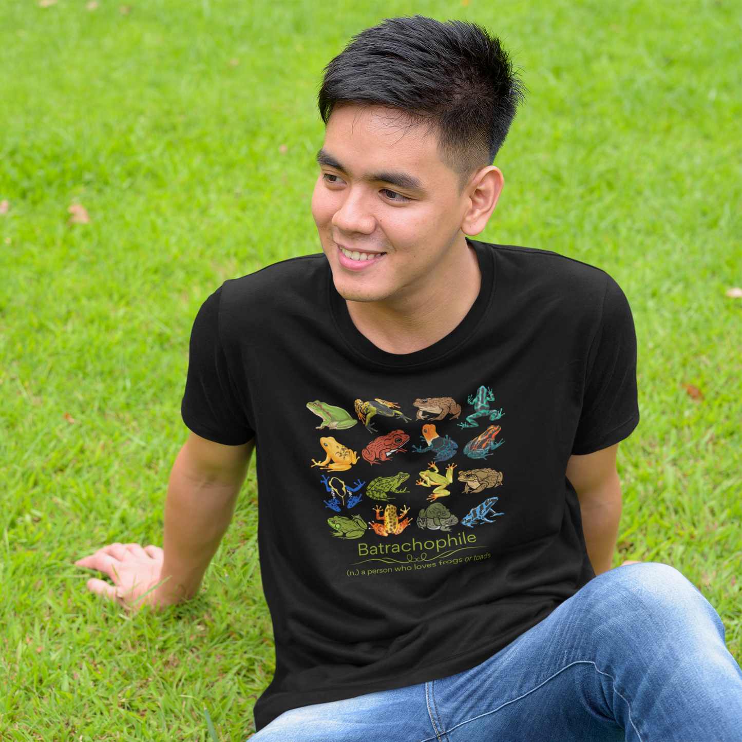Young Asian man relaxing on the grass wearing jeans and a black t-shirt with 16 colorful frogs and toads in a 4x4 pattern; says Batrachophile - a person who loves frogs or toads below the frog images.