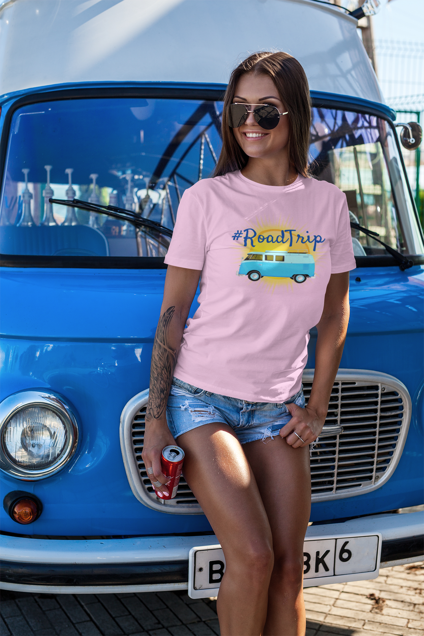 Attractive woman leaning up against a blue van wearing shorts and a pink t-shirt that has a similar van on the front with a sun behind it and a hashtag message reading #RoadTrip