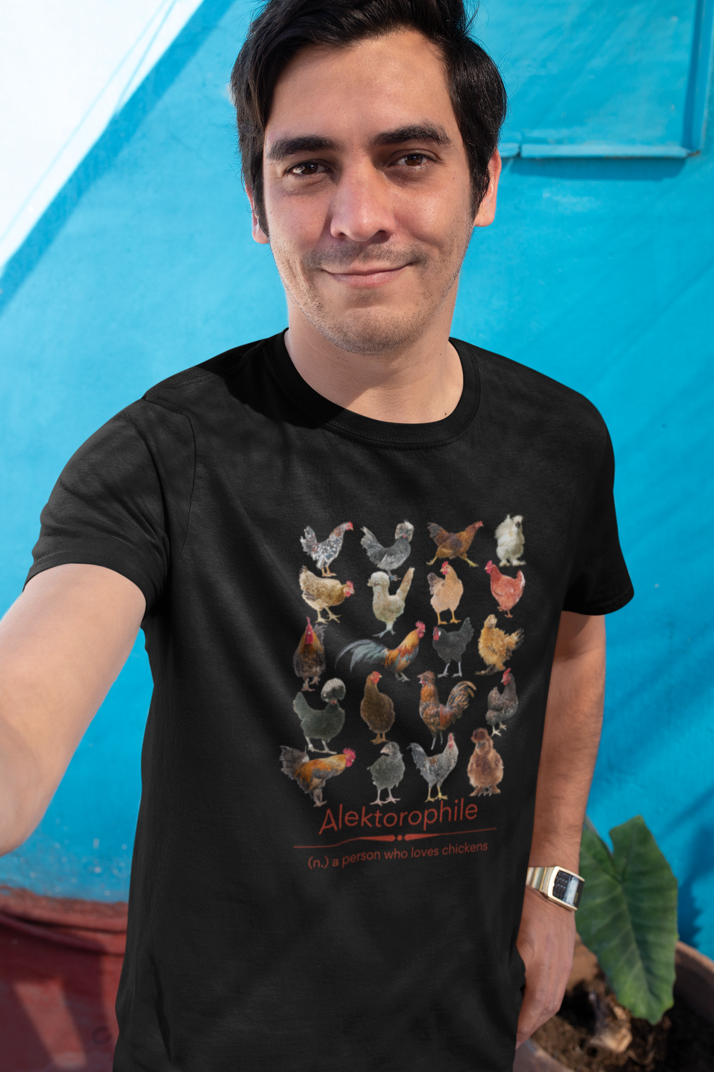 Smiling Latino man taking a selfie wearing a black t-shirt with 20 colorful chickens in a 4x5 pattern and underneath says 'Alektorophile - a person who loves chickens' silkies, bantams, polish, and many more.
