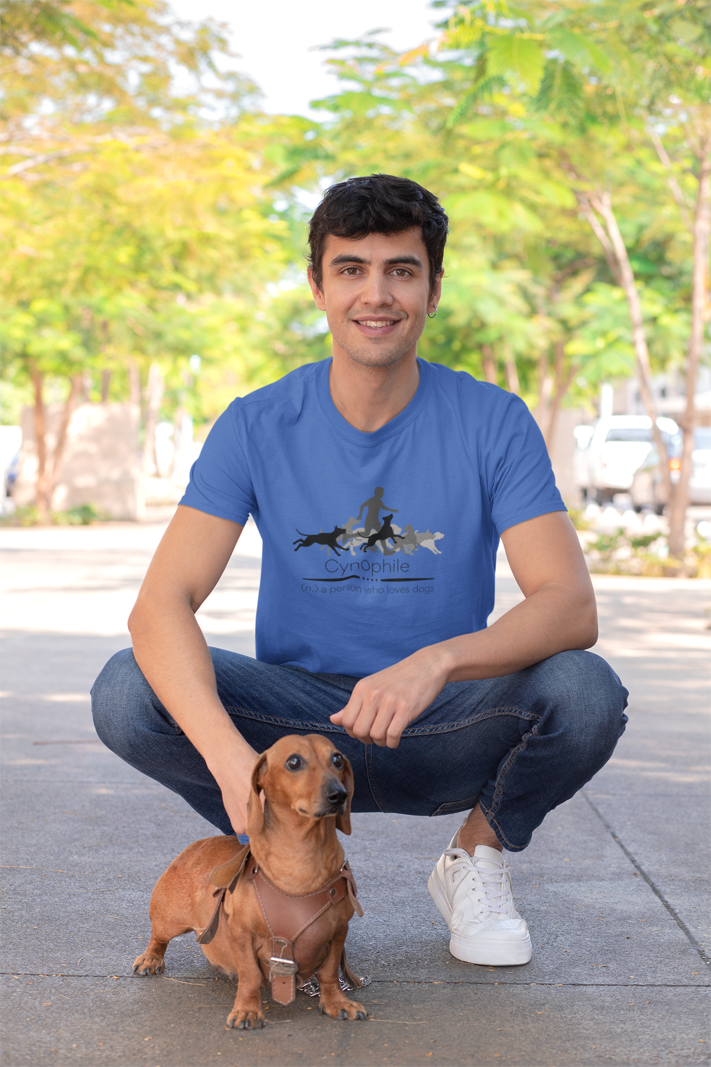 Brunette man kneeling wearing jeans and blue t-shirt designed with silhouette of a man with several dogs and also says 'cyanophile - a person who loves dogs'; great gift for a dog lover