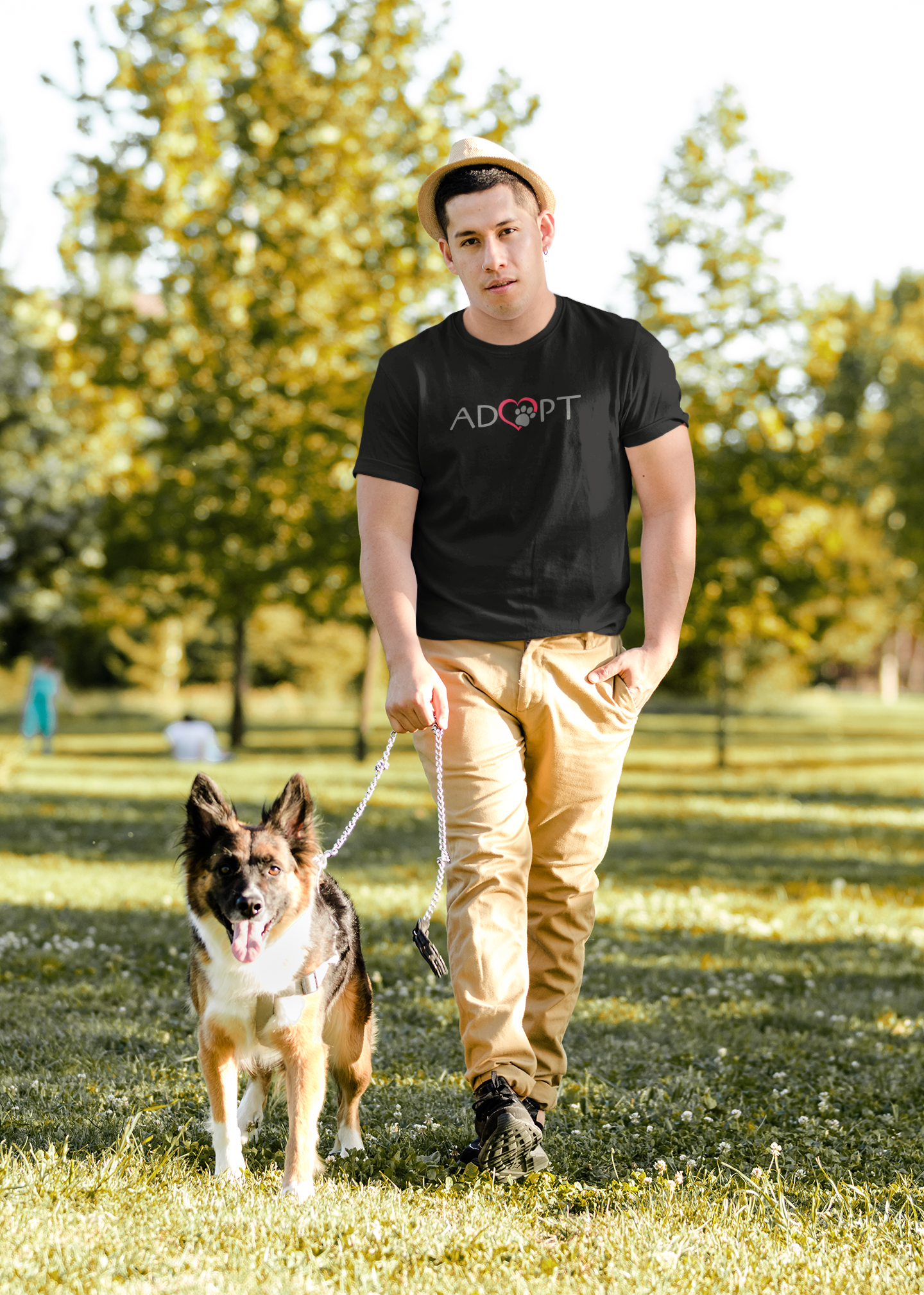 A handsome young man walking his dog in a grassy setting with trees; the man is wearing a black t-shirt that says ADOPT on it but the letter O is the shape of a red heart with a paw print in the center to promote adopting pets